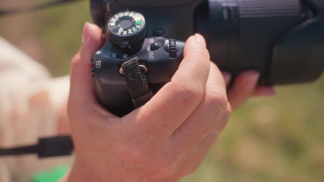 Close up view of fair skin hand adjusting DSLR camera settings with dial in natural light outdoors, lens in focus with soft blurred green background