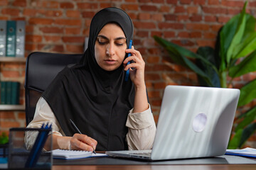 Middle Eastern Muslim woman in black hijab working at a desk in a loft style office.
