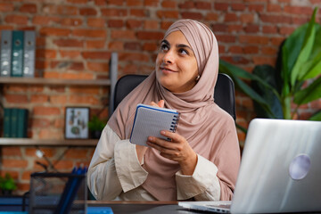 Contemplative Woman in Hijab Holding Notebook at Office Desk
