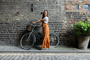 Smiling woman with bicycle walking by brick wall in urban city street