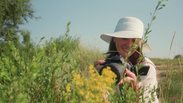 Smiling woman in white sunhat squats joyfully among green wild plants holding camera while taking photo of yellow flowers in sunny meadow surrounded by tall grasses under clear blue sky