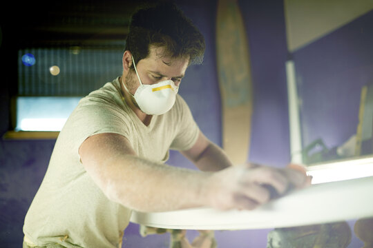 Craft, surfboard and polish with man in workshop for manufacturing, sports equipment and craftsman. Entrepreneur, maintenance and repair with person and mask for small business and quality assurance - Powered by Adobe