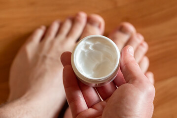 Person holds cream with bare feet in background. Hands holding a cream tube with bare feet in the background, preparing for moisturizing