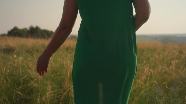 close up rear view of woman in green dress walking through tall dry grass during warm daylight moment, softly brushing fingertips along field stems in peaceful countryside setting