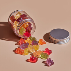 Open transparent jar of colorful gummy bears spilling onto a soft pastel surface, with the silver lid placed nearby, creating a playful and vibrant composition.