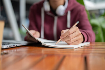 Close up college student man writing in notebook with a pencil aside laptop sitting at table in cafe