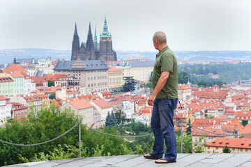 Mature tourist poses proudly above red rooftops of Prague, with iconic cathedral behind