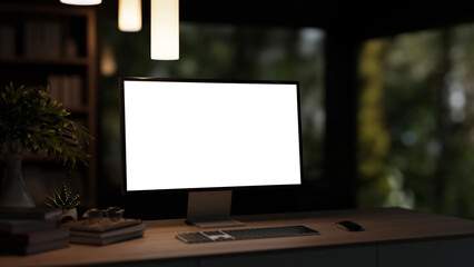 Fototapeta premium Blank screen computer with books aside pot plants on wooden table under hanging lamp in dim lit room