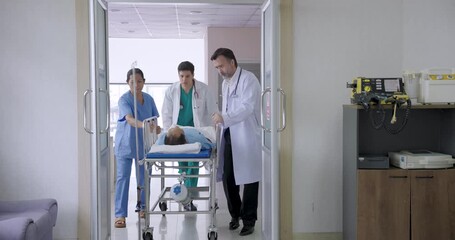 emergency medical team including caucasian and asian doctors and nurse pushing unconscious male patient on stretcher through hospital hallway toward operating room for immediate trauma treatment