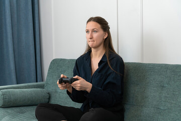 Young woman sitting on a comfortable sofa holding a video game controller while playing videogames, concentrating on the screen with a serious expression