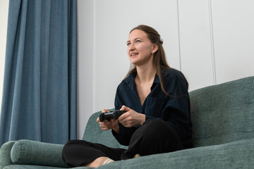 Young woman sitting comfortably on a vibrant green sofa in her living room, holding a gamepad and fully immersed in playing video games, relishing her leisure time and enjoying every moment