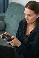 Young woman sitting on a comfortable sofa playing videogames with a wireless controller, concentrating on the game with a serious expression. Vertical photo