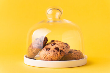 Stack of fresh chocolate American style chip cookies with nuts and chocolate drops on a tray with transparent lid on yellow background. Cookies with peanut butter filling inside a glass jar