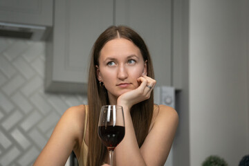 Thoughtful young woman sitting at a kitchen counter, holding a glass of red wine and gazing into the distance, immersed in deep contemplation and reflection on life's challenges