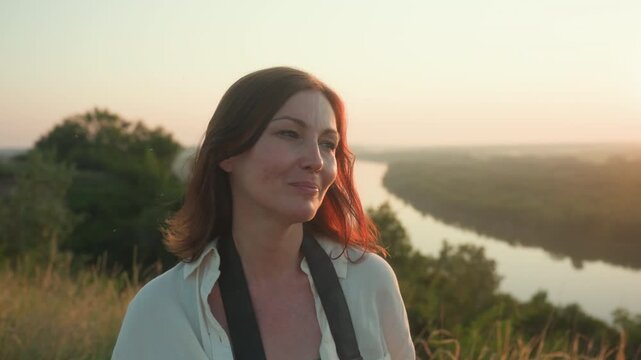 Lady with subtle expression faking smile under warm sunset light in tall grassy field, soft golden rays highlighting hair as river and trees blur in distant background
