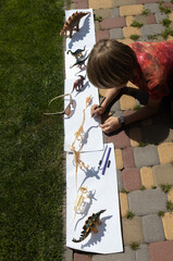 boy lies on paving slab and traces contours of contrasting shadows from figures of toy dinosaurs on...