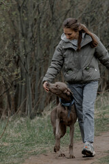 A young Caucasian woman with brown hair runs joyfully on a dirt path with a playful brown dog in a green, wooded area.