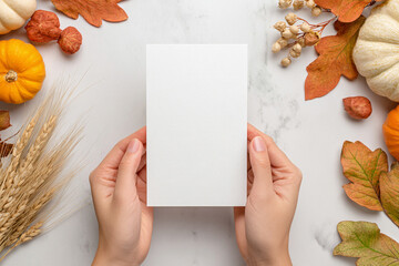 Two hands hold a blank greeting card for Thanksgiving invitation mockup amid a vibrant autumn display featuring pumpkins, leaves, and wheat