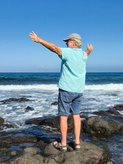 Rear view of a senior man with outstretched arms enjoying sunny day by the sea looking at the...