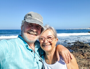 Happy senior couple looking at camera embracing while enjoying sunny day by the sea in summer environment, active retirement, leisure and vacation