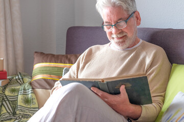 An elderly man with a white beard and hair relaxes at home, reading a book, comfortably seated on...