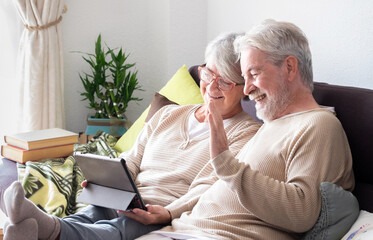 A modern, smiling older couple relaxes at home, sitting comfortably on the couch, surfing the...