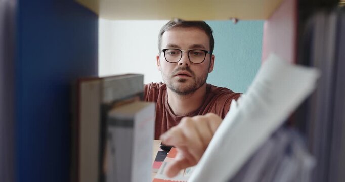 A young man browses through documents and binders on a shelf, clearly searching for something important. His expression shifts to disappointment as he fails to find what he was looking for.