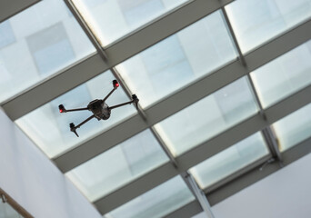 A drone hovers beneath a glass ceiling within a contemporary structure, utilizing bright red lights as it navigates the interior space during daylight hours.