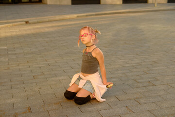 Pensive young girl in pink glasses kneeling on pavement outdoors at sunset, wearing black top, denim shorts, and light jacket tied around waist, capturing a reflective childhood moment
