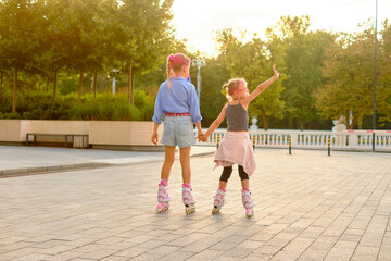 Joyful sisters in pink glasses roller skating outdoors in city park at sunset, holding hands and sharing smiles during lively family activity