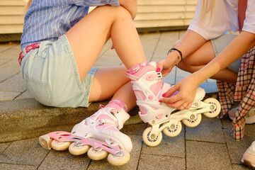 Young woman helping girl with pinkroller skates sit on city sidewalk during sunny day, capturing outdoor friendship, active lifestyle, joyful childhood moments