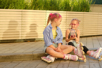 Two young girls with colorful braided hair and roller skates sitting by modern wooden fence on sidewalk, sharing a refreshing berry drink during sunny day outdoor leisure