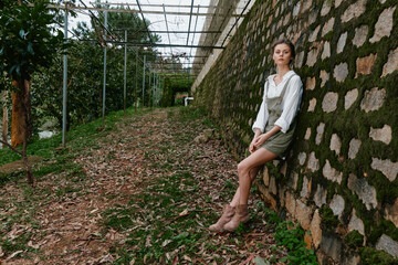 Young woman in casual outfit sitting on stone wall surrounded by greenery, conveying a serene and...