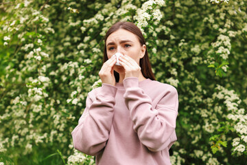 Woman with paper tissue suffering from seasonal pollen allergy near blossoming shrub on spring day