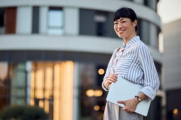 Confident Asian Businesswoman Standing Outdoors Holding a Laptop Near Modern Office Building