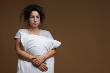 Woman with eye patches and pillow suffering from hangover on brown background, space for text
