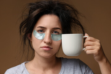 Woman with eye patches and cup of drink suffering from hangover on brown background