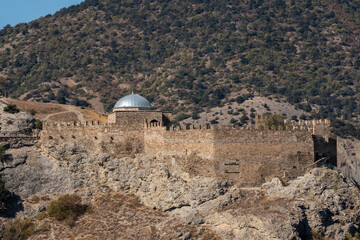 Fragment of Genoese fortress wall stands on top of rocky cape Alchak, surrounded by rugged terrain and sparse vegetation against natural backdrop. Sudak, Crimea