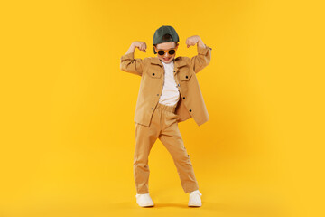 Happy little boy in sunglasses and baseball hat dancing on orange background