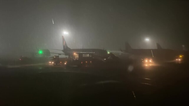 Night Airport in Heavy Fog. Atmospheric nighttime footage of a busy airport during heavy rain and fog. Silhouettes of airplanes and special equipment with flashing beacons in the light of searchlights