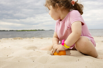  A small girl sitting peacefully on the riverbank, holding a bright orange in her hands, surrounded...