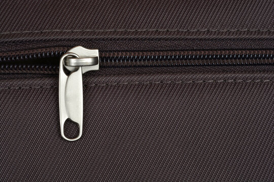 A close-up shot of a silver zipper pull on dark brown textile, revealing its metallic texture and the fine stitching around the opening - Powered by Adobe