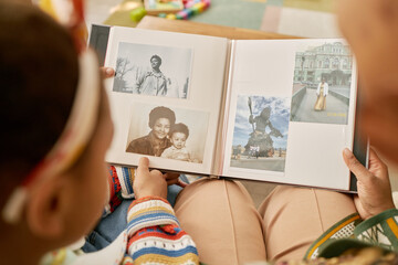 Over shoulder shot of Black woman with daughter looking at family photo album with diverse pictures from past, connecting through familial heritage at home