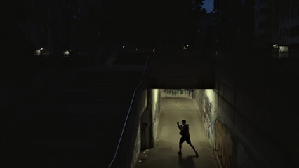 A lone figure in a hoodie and shorts practices boxing in a dimly lit, graffiti-covered underpass, embodying resilience and determination amidst urban grit, captured with a wide shot.