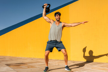Hispanic man performs kettlebell exercise outdoors in vibrant urban setting under bright blue sky