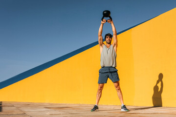 Hispanic man performs kettlebell workout outdoors against vibrant colorful wall in bright, sunny...