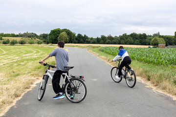 Two boys cycling on a quiet rural road surrounded by green fields and trees.
