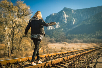 Happy blonde girl balancing walking on railway track with arms spread in mountain landscape. Girl balancing on train rails in scenic landscape with rocky cliffs and autumn trees outdoors.