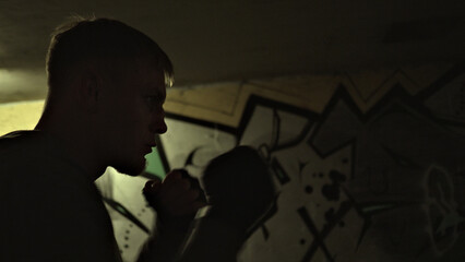 Silhouette of a focused male boxer in a dimly lit urban setting with graffiti, embodying resilience and determination. His intense gaze and poised stance highlight his warrior spirit amidst.