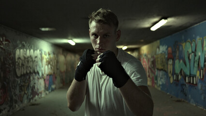 A determined Caucasian male boxer stands in a graffiti-covered alley, exuding resilience and strength. His intense gaze and poised stance capture the essence of combat sport.